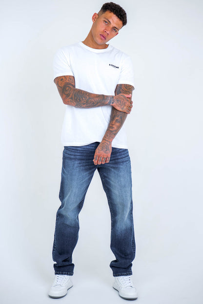 A man with tattoos wears a white t-shirt, B Couture Beckenham Straight Fit Stretch Denim Jeans in dark blue, and white sneakers. He stands against a plain light background with one arm crossed over his chest, looking at the camera.