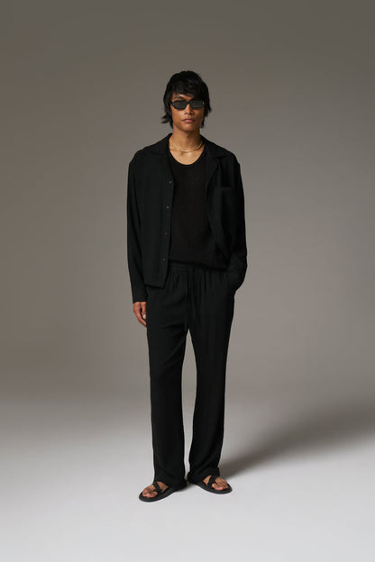 A man wearing a long sleeved shirt, with black sunglasses and pants against a neutral background.